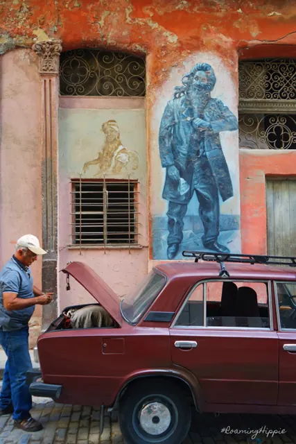 A man working under the hood of an old red car beside a painted mural