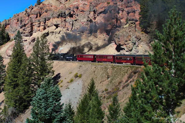 A red passenger train curving along a rocky cliffside through pines
