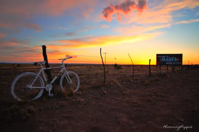 A white bicycle by a fence at sunset with a Marfa billboard behind it